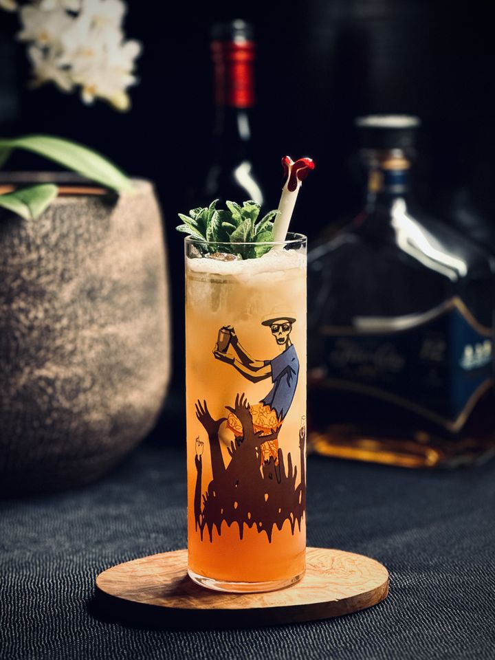 a brown cocktail in a Beachbum Berry 1934 Zombie glass with a mint bouquet garnish and glass Surfside Sips bloody bone straw. In the background are white orchids and bottles of rum