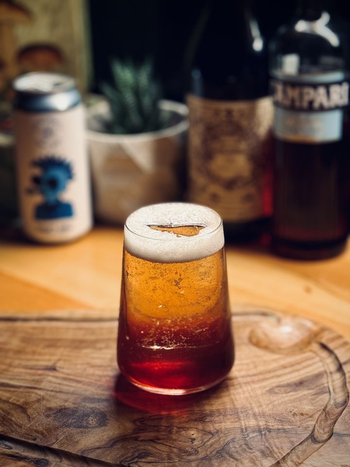 a yellow to dark red gradient colored cocktail with a long spear of clear ice sitting on an olive wood cutting board with cans and bottles in the background: Les Intenables Crispy Chill hoppy lager, Carpano Antica, and Campari