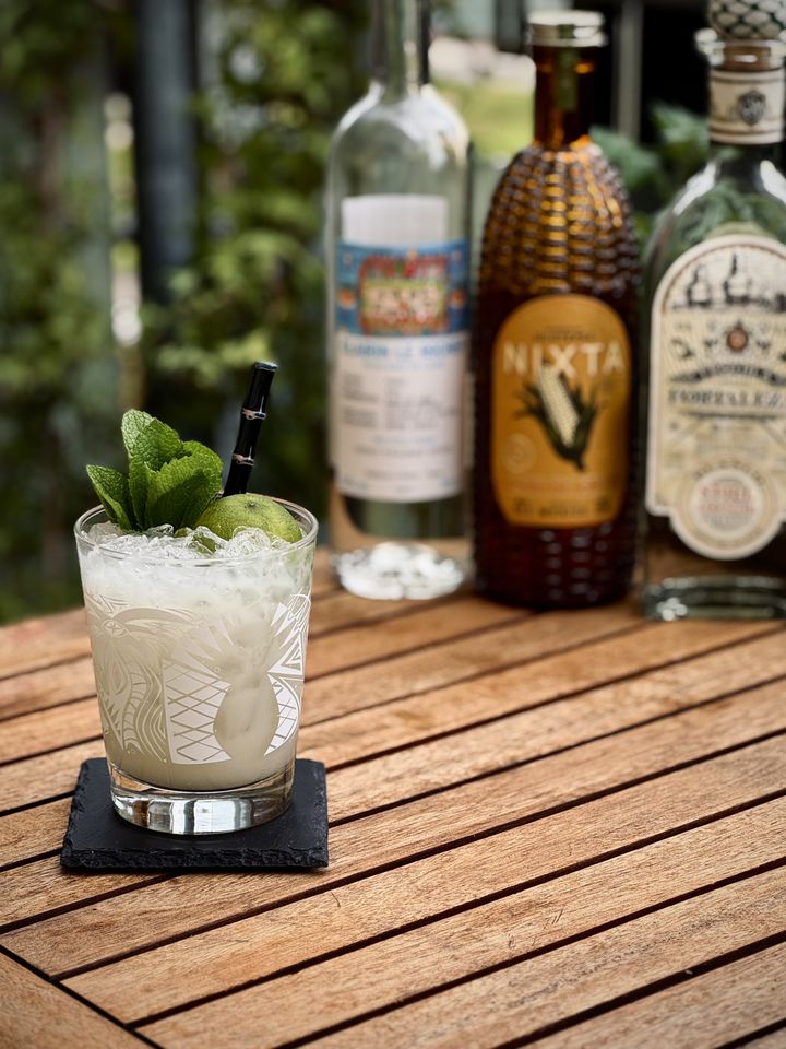 a milky white cocktail with mint bouquet and spent lime shell garnish placed in a B-Rex Mai Tai glass with a Surfside Sips realistic black bamboo straw. The drink is on a wooden table with bottles of tequila, Nixta Licor de Elote, and Clairin in the background