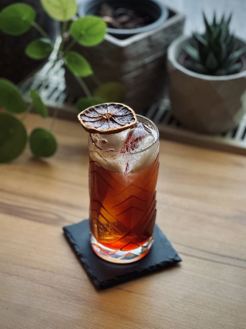 a reddish brown cocktail with long spear of clear ice and dehydrated blood orange garnish sitting on a black slate coaster on a wooden table with potted plants in the background