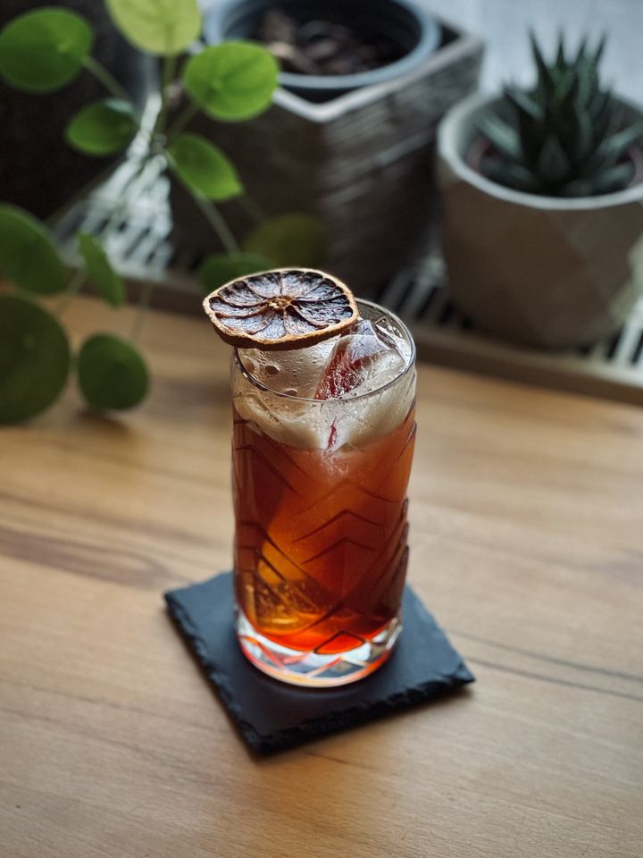 a reddish brown cocktail with long spear of clear ice and dehydrated blood orange garnish sitting on a black slate coaster on a wooden table with potted plants in the background