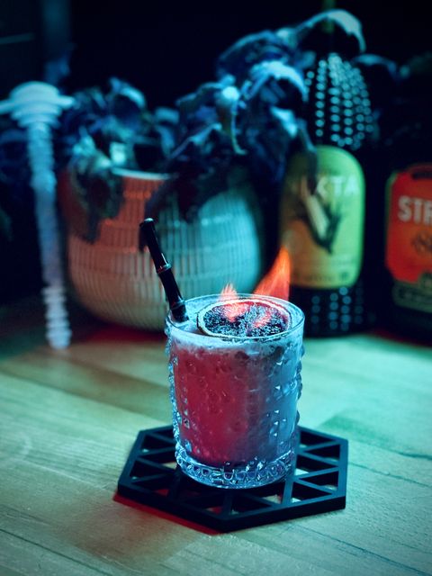an off-white cocktail lit by two colored lights, turquoise and pink, with a flaming dehydrated blood orange garnish and a Surfside Sips black realistic bamboo glass straw sitting on a hexagonal coaster with a background consisting of a weird potted plant, bottles of Stroh 80, Nixta Elote, and a Mothership swizzle stick