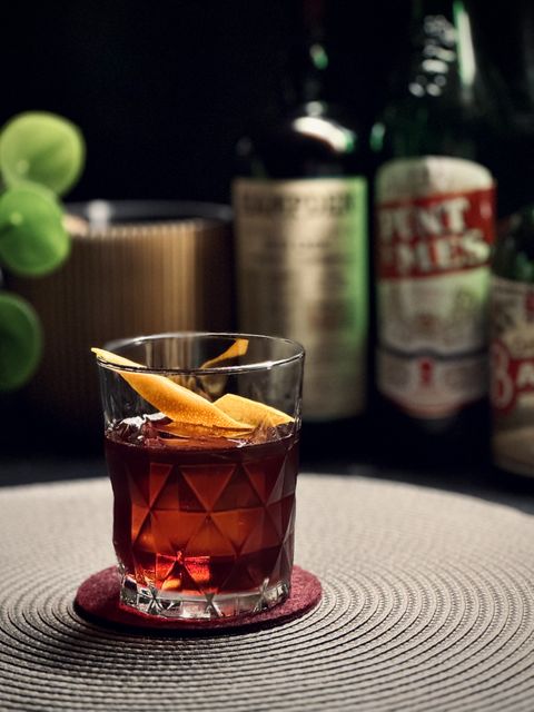 a deep red cocktail with big rock of clear ice and orange peel garnish sitting on a burgundy felt coaster. In the background are bottles of rum and amaro