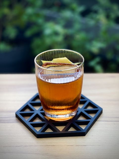 a brown cocktail with big rock of clear ice and lemon peel garnish sitting on a black silicone coaster on a wooden table with plants in the background