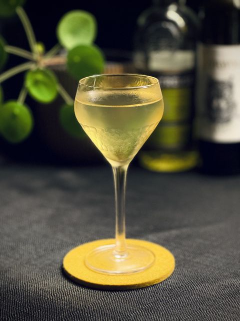 a clear yellow cocktail in a chilled coupe glass on a yellow felt coaster. In the background is a potted plant and bottles of yellow Chartreuse and vermouth