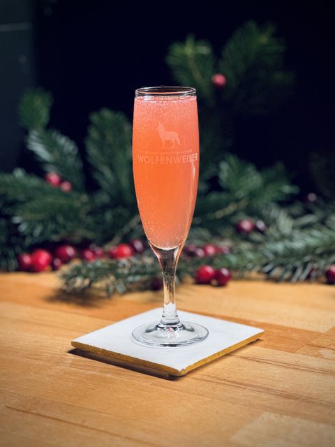 bright pink bubbly cocktail in a tall Wolfenweiler sekt glass sitting on a white coaster with pine tree branches and cranberries in the background
