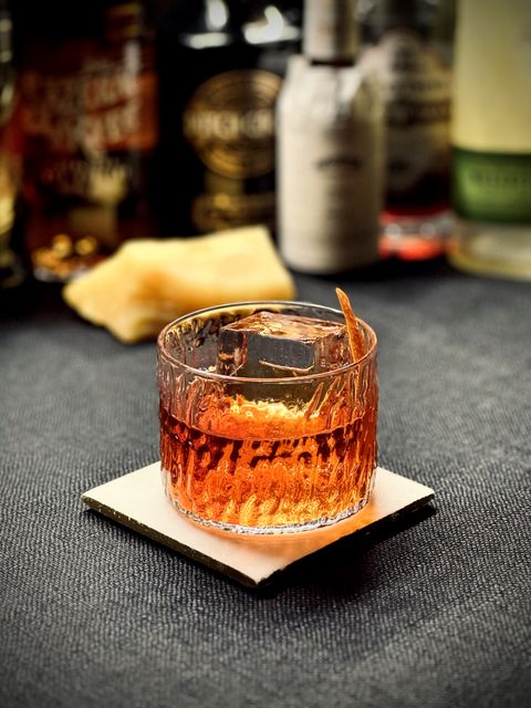 a deep orange or light brown cocktail with a big rock of clear ice and orange peel garnish sitting on a white coaster in front of bottles and a chunk of beeswax