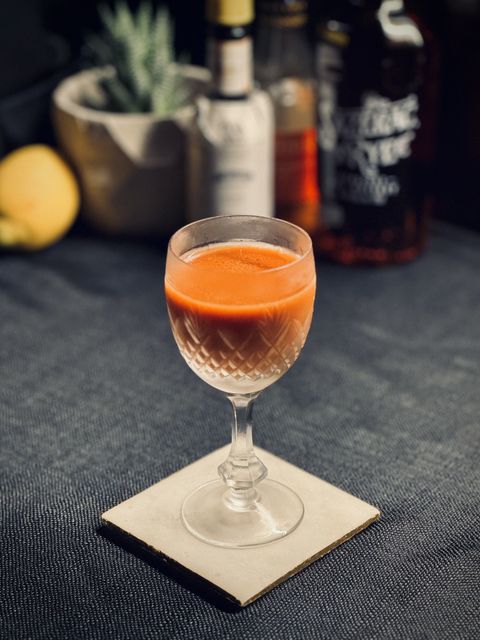 a bright orange cocktail in a frosty coupe glass with bottles of Angostura bitters, Liber & Co orgeat and Sazerac rye whiskey, and a lemon in the background