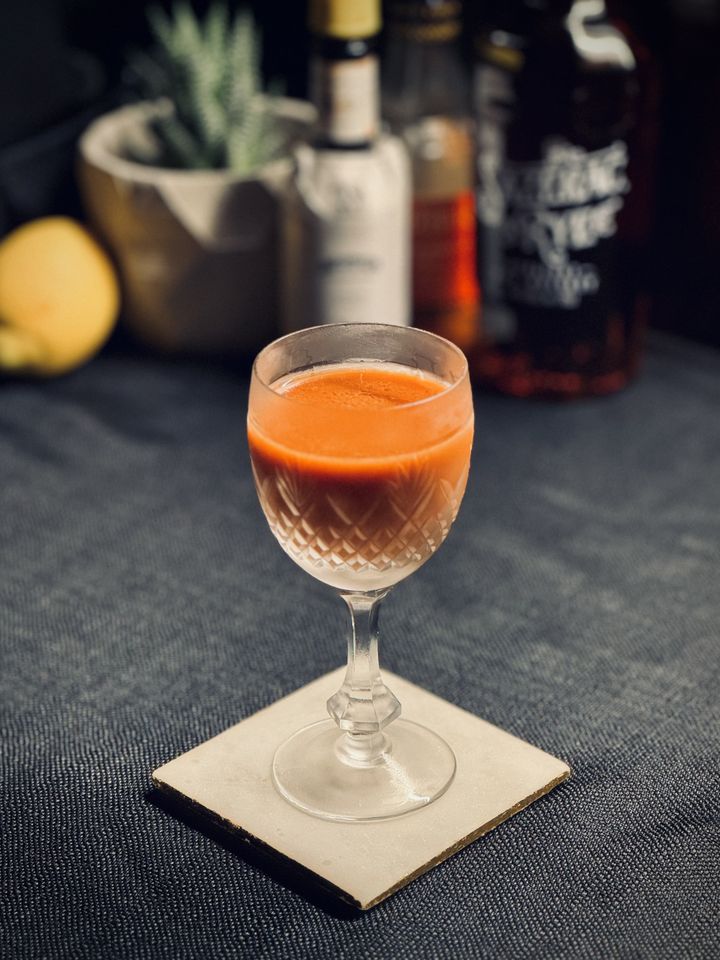 a bright orange cocktail in a frosty coupe glass with bottles of Angostura bitters, Liber & Co orgeat and Sazerac rye whiskey, and a lemon in the background