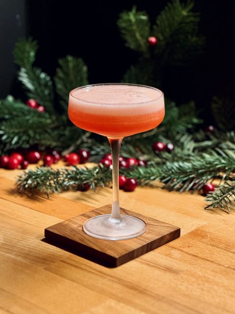 a bright red cocktail with bubbly head in a chilled coupe sitting on a wooden coaster with pine branches and cranberries in the background