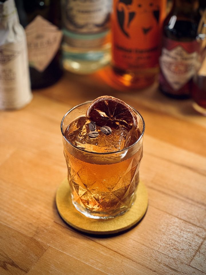 a golden yellow cocktail with big rock of clear ice, Nomad coffee beans, and dehydrated orange slice garnish sitting on a yellow felt coaster with bottles in the background