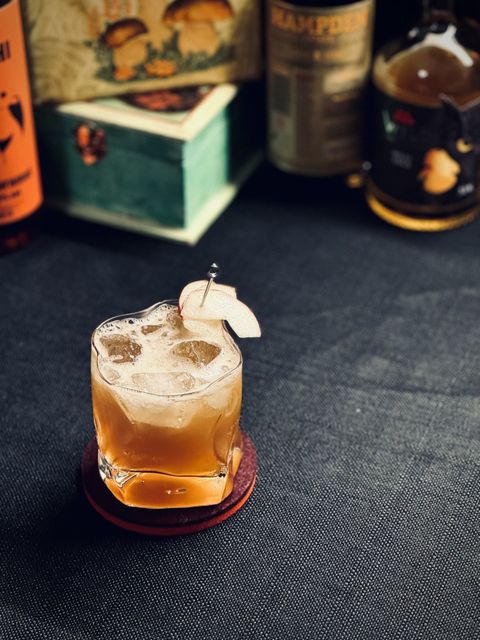 a brown cocktail with fresh pear slice garnish in a wobbly warped glass. in the background are bottles of rum and liqueur