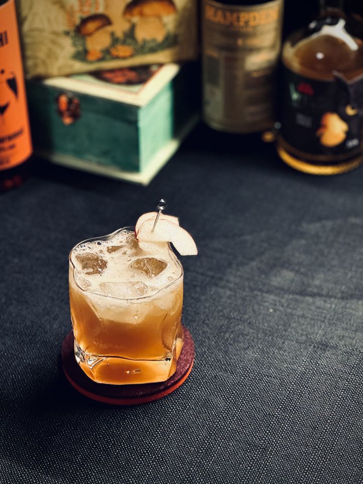 a brown cocktail with fresh pear slice garnish in a wobbly warped glass. in the background are bottles of rum and liqueur