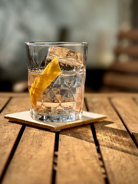 crystal clear slightly pink cocktail with lemon peel garnish and large clear ice cube sitting on a white coaster and wooden table with sunshine caustics on the table