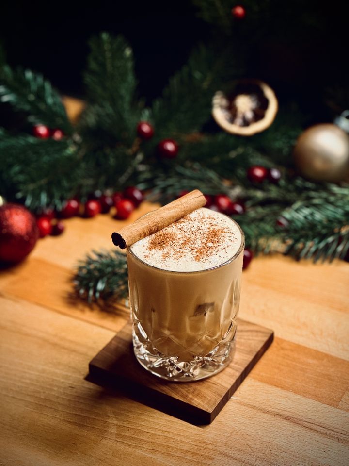 a light brown creamy cocktail with grated nutmeg and cinnamon stick garnish sitting on a coaster in front of pine tree branches, cranberries, christmas tree ornaments, and dehydrated orange slices