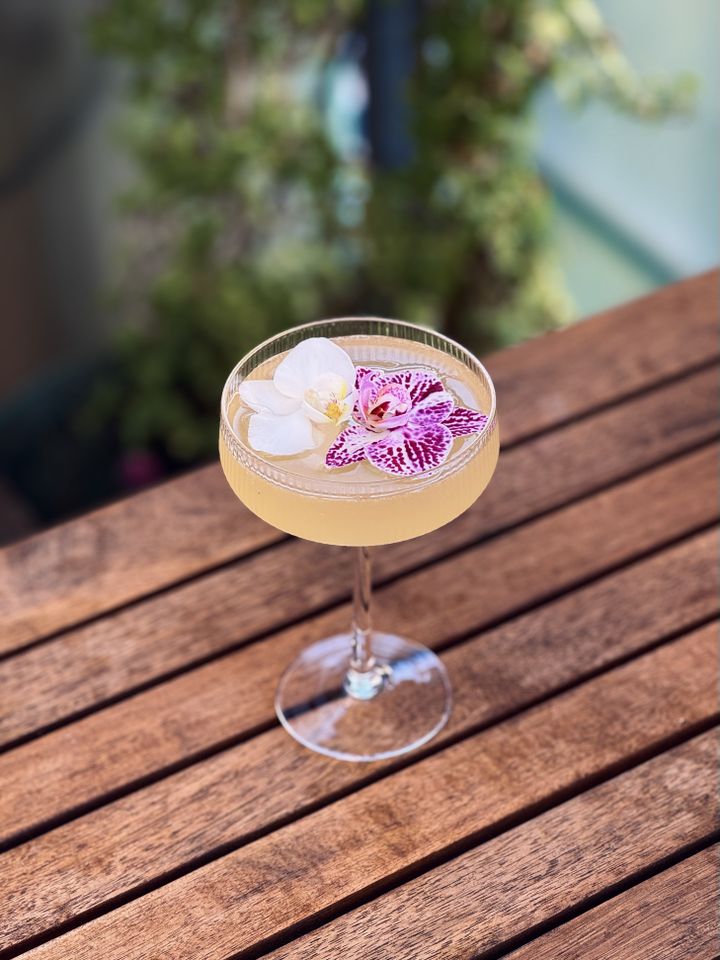 light yellow cocktail with both white and speckled purple orchid garnish in a coupe glass sitting on a wooden table with plants in the background