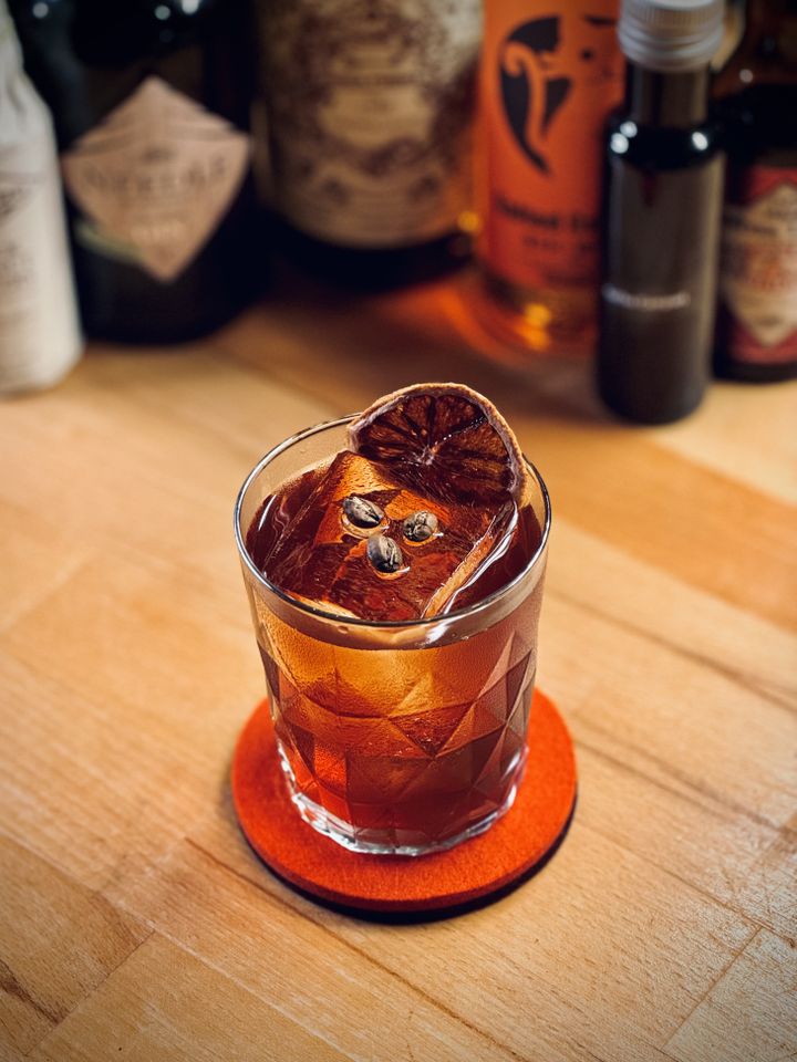 a dark red cocktail with big rock of clear ice, three coffee beans, and a dehydrated orange slice as garnish sitting on a red felt coaster with bottles in the background