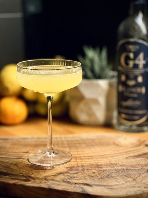 a bright yellow cocktail i garnished in a coupe glass sitting on a wooden cutting board with fruit, plants and a bottle of G4 still strength tequila blanco in the background