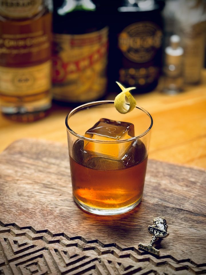 a dark amber brown cocktail with crystal clear big rock of ice and lemon twist garnish sitting next to a Youngblood cocktail bar pin on a wooden cutting board with bottles in the background