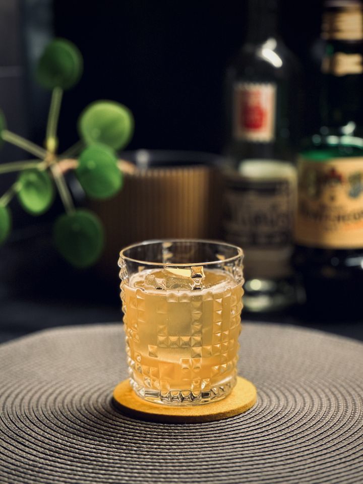 a golden yellow cocktail with big rock of clear ice an fresh ginger slice garnish in a Nachtmann crystal glass sitting on a yellow felt coaster. In the background are bottles of mezcal and Amaro Montenegro