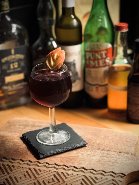 a dark brown cocktail with carved apple garnish in a coupe glass sitting on a cutting board with bottles in the background