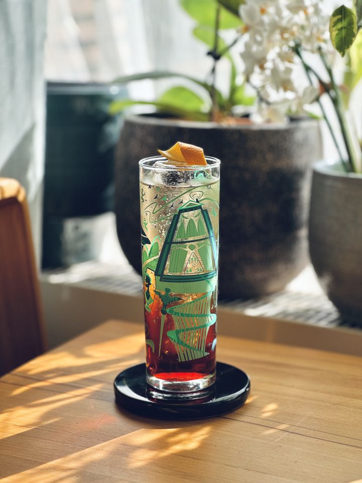 an unstirred cocktail half clear and half dark red with a long spear of clear ice and orange twist garnish sitting on a wooden table with sunlight shining through leaves