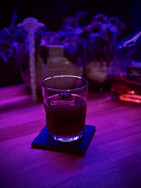 a dark, cloudy cocktail with big rock of clear ice and star anise garnish, sitting in a colorfully lit setting with a strange potted plant, bottles of rum, and a Mothership swizzle stick in the background