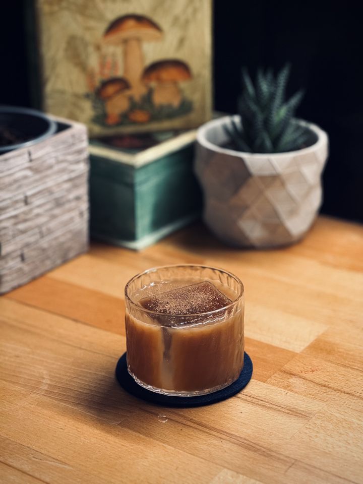 a cloudy brown cocktail with grated nutmeg and a big rock of clear ice sitting on a wooden countertop with plants in the background