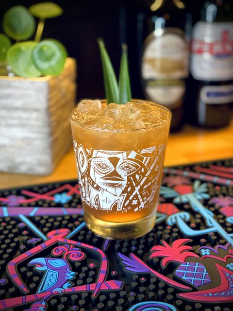 brown colored cocktail with pineapple frond garnish in a B-Rex mai tai glass sitting on a B-Rex pineapple bar mat with a bottle of Hamilton Pot Still Black rum, a bottle of Pernod, and a potted plant in the background