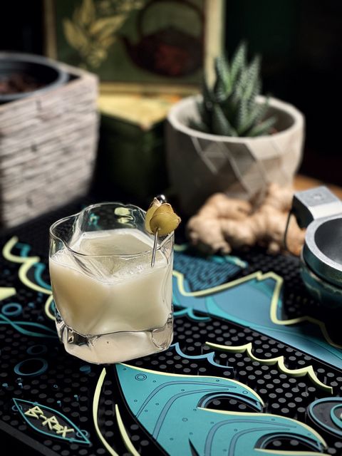 a milky white cocktail with big rock of clear ice and fresh ginger garnish in a wavy old fashioned glass sitting on a B-Rex bar mat with a lime squeezer, a root of ginger, and potted plants in the background