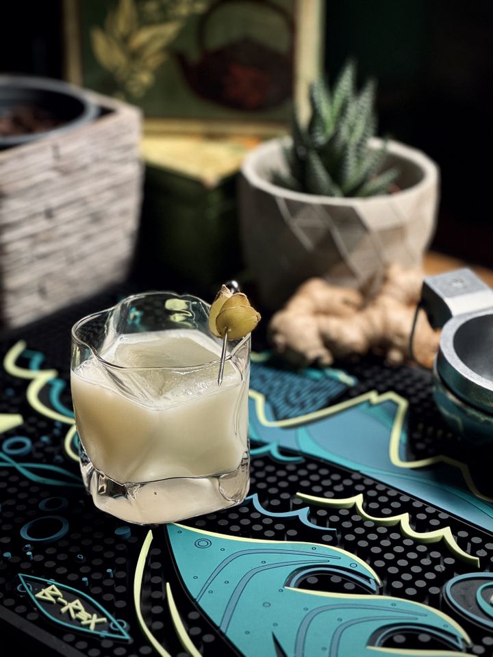 a milky white cocktail with big rock of clear ice and fresh ginger garnish in a wavy old fashioned glass sitting on a B-Rex bar mat with a lime squeezer, a root of ginger, and potted plants in the background