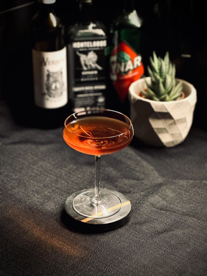 a dark red cocktail in a coupe glass with bottles and a plant in the background