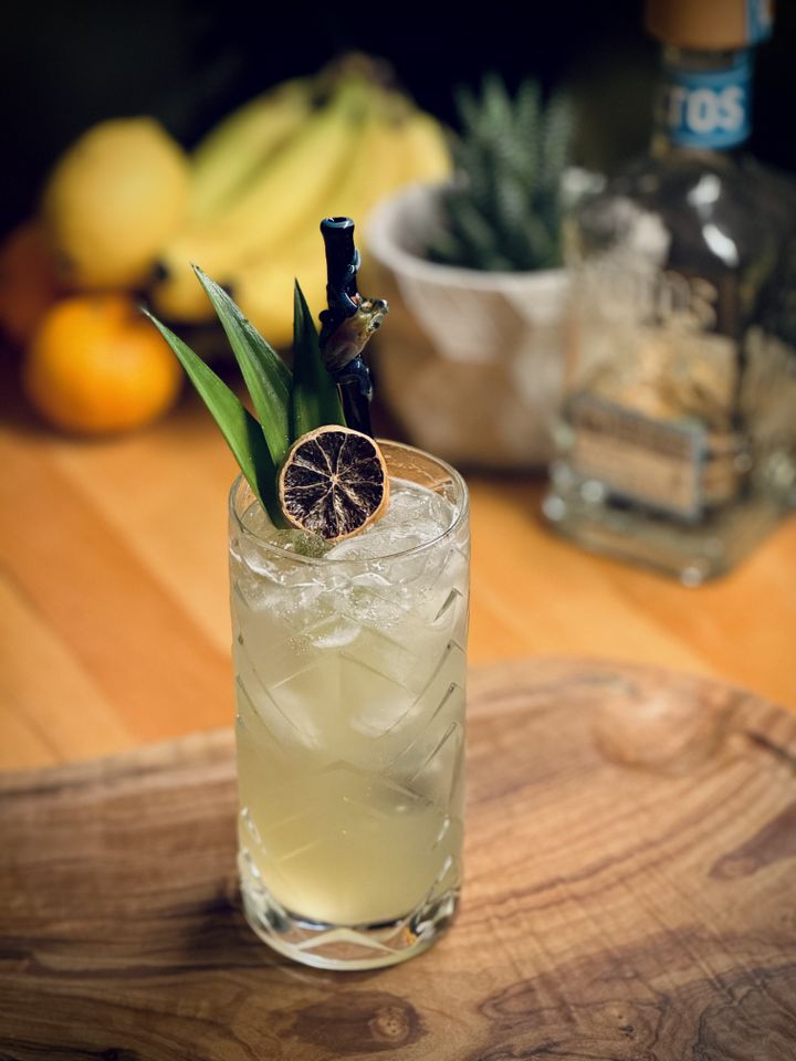 a light yellow colored cocktail with pineapple frond garnish and surfside sips realistic glass bamboo straw sitting in front of fruit, plants, and a bottle of Olmeca Altos tequila blanco