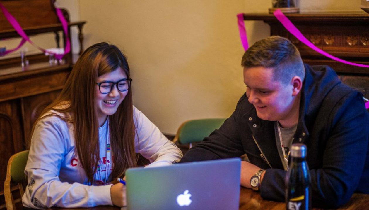 Two students sit together at a table, smiling and looking at a laptop.