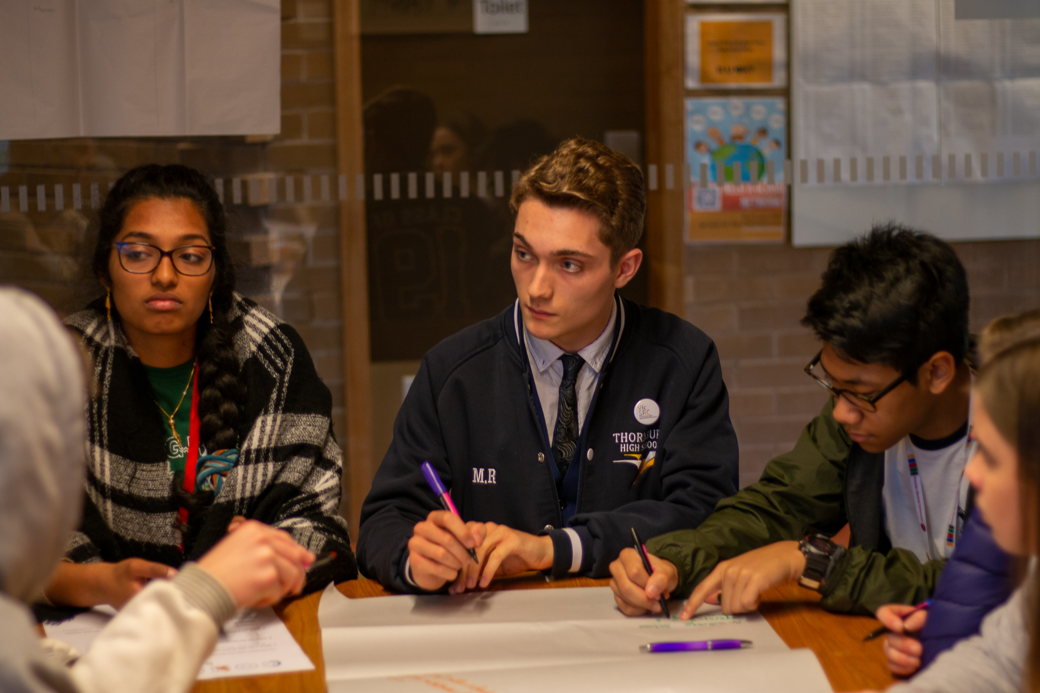 Students sit at a table, deep in conversation. Three students are clearly visible but there are more students sitting closer to the camera. The student in the centre of the three is listening intently, clearly ready to write something down, while another student beside him is already writing.