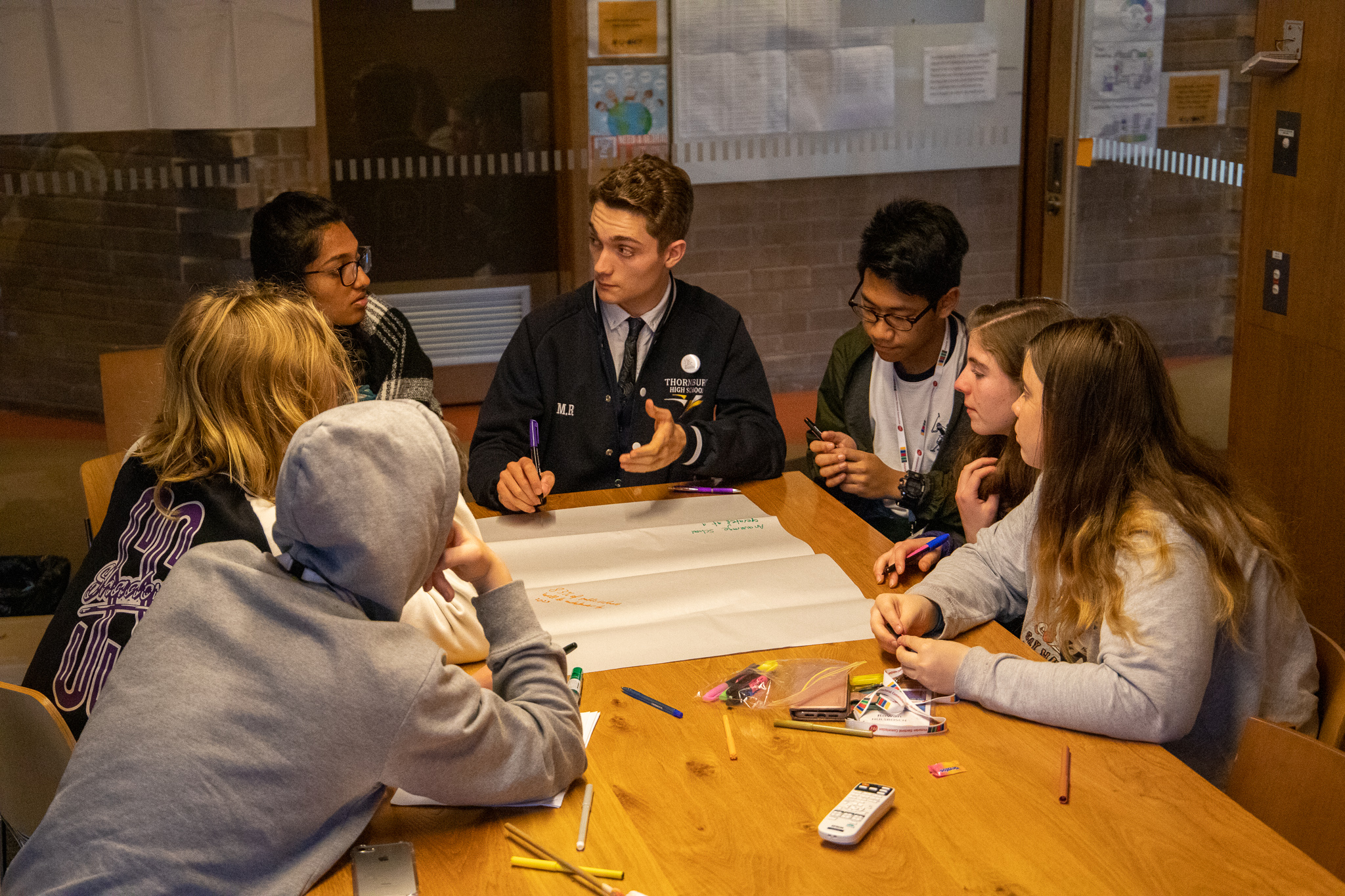 A group of seven students are sitting around a wooden desk. They are talking animatedly and on the desk is a piece of butchers paper and lots of pens and markers.