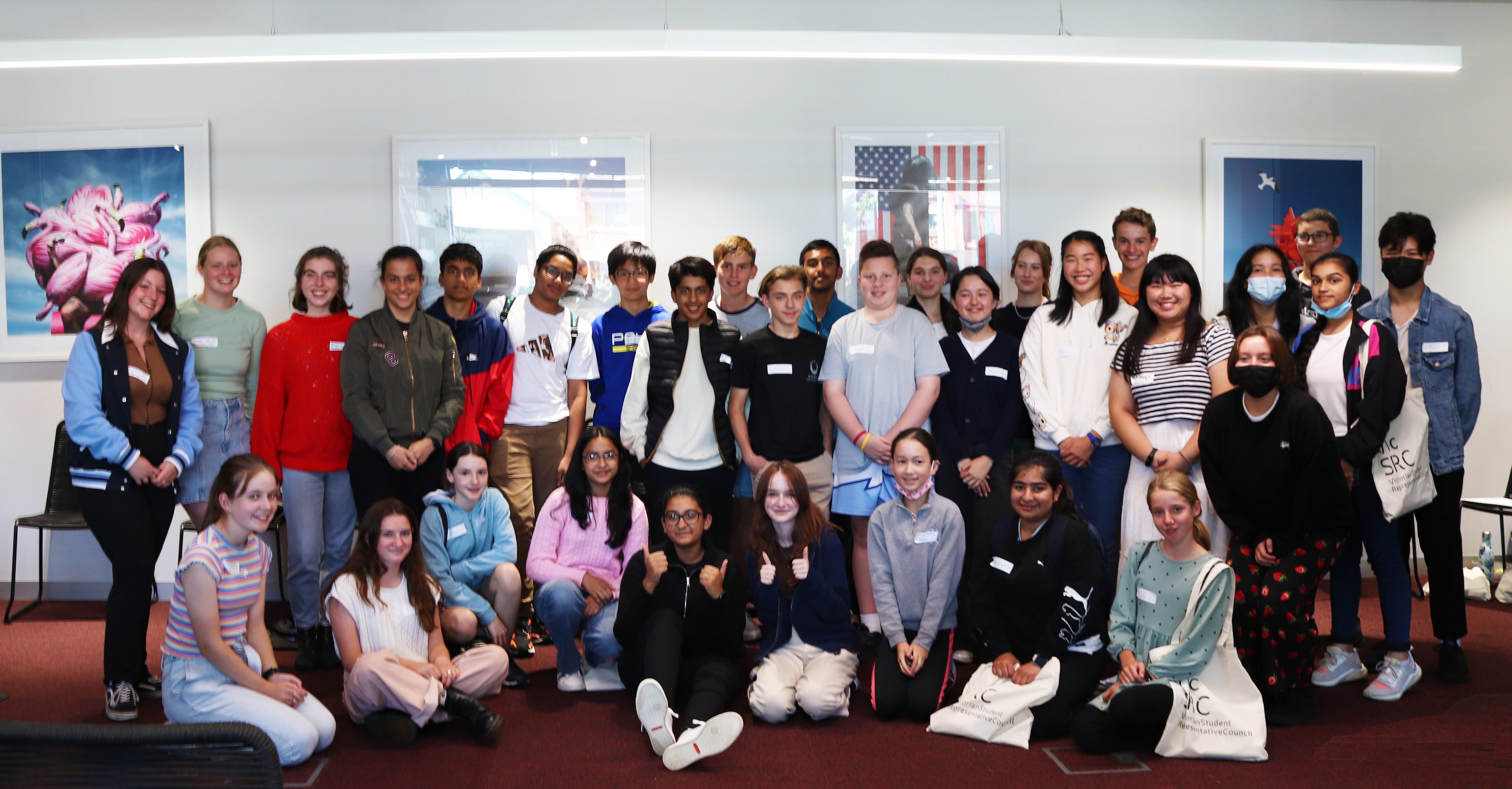 A group of 30 young people in casual clothes are standing together. There are paintings on the wall behind them and they are all smiling at the camera.