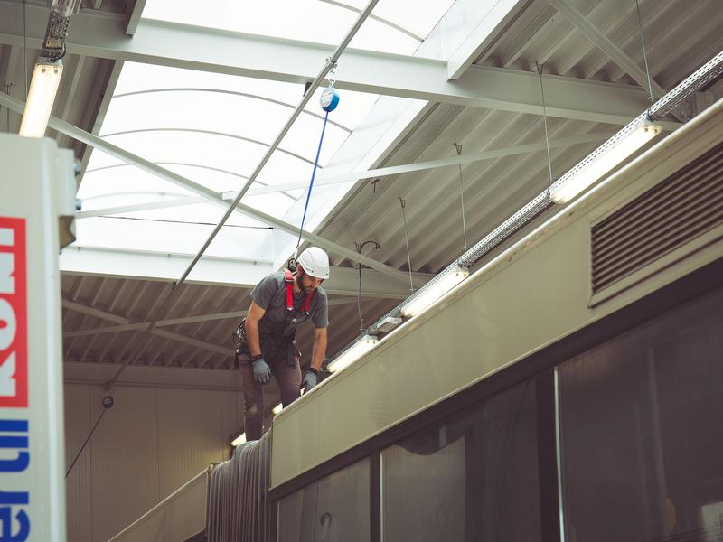 Werker met harnas op een voertuig in een hal, met een valstopapparaat aan een rail erboven