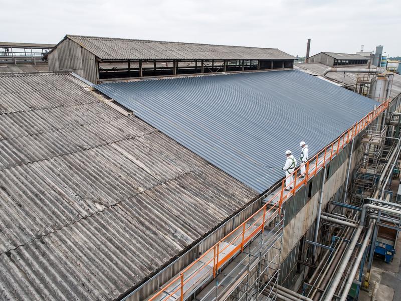 Oud dak van golfplaten naast nieuwe metalen dakplaten op een fabriekshal, met twee werkers in beschermpak op een loopbrug