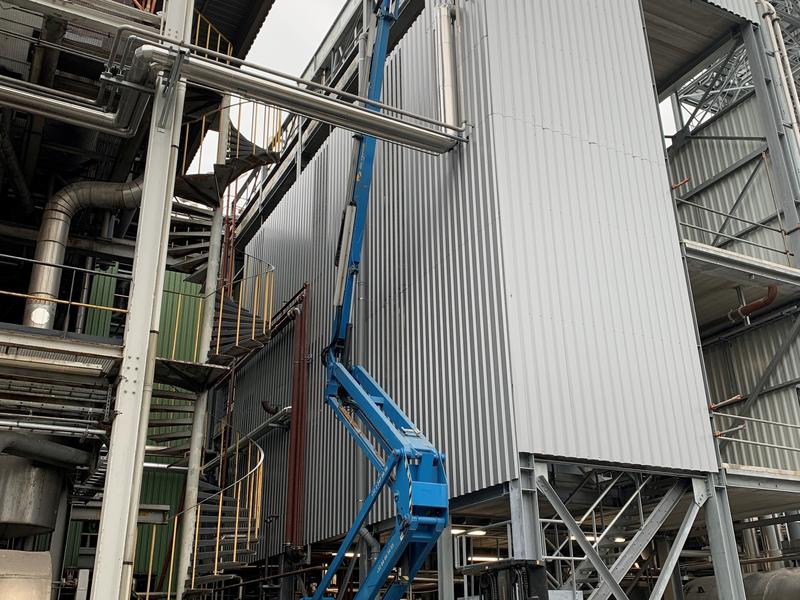 Grijze geprofileerde metalen panelen op een industriële installatie, met een blauwe hoogwerker ervoor