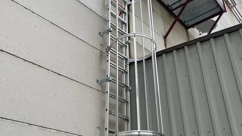 Aluminium kooiladder tegen een gevel van betonplaten, naast een rood stalen bordes
