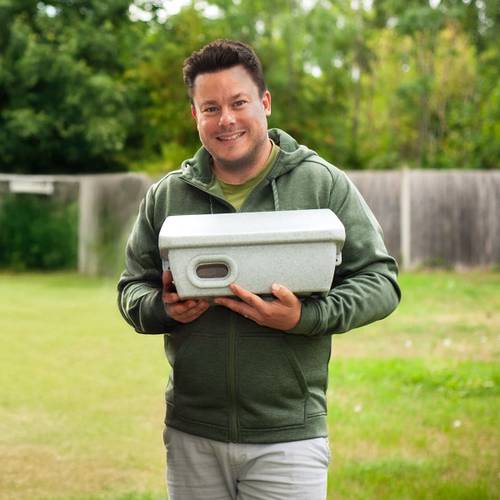 Man in a green hoodie smiles while holding a grey rectangular object outdoors, with trees and a fence in the background.