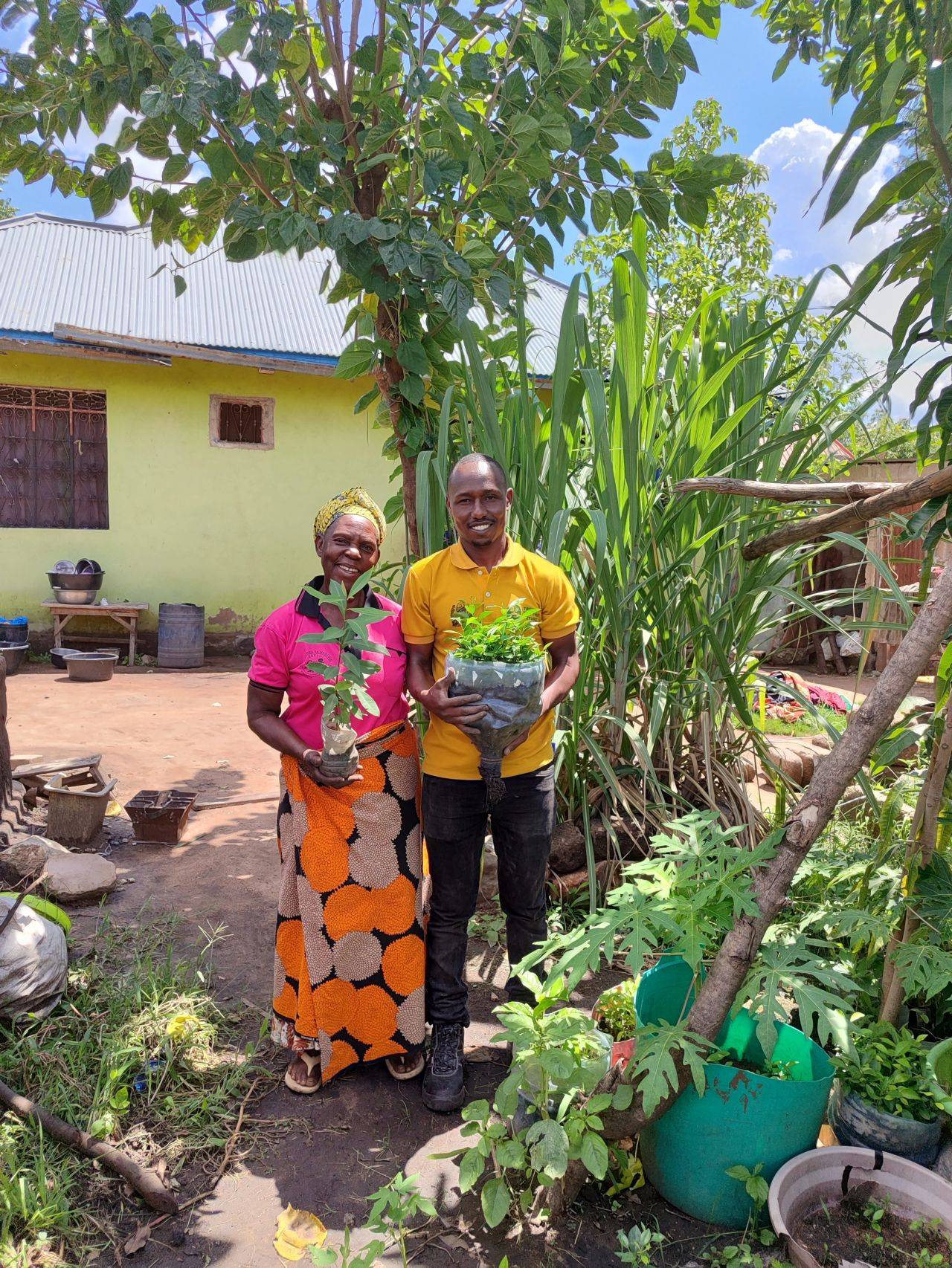 Two people standing in a garden, holding young plants in containers, surrounded by lush greenery and a yellow house in the background.
