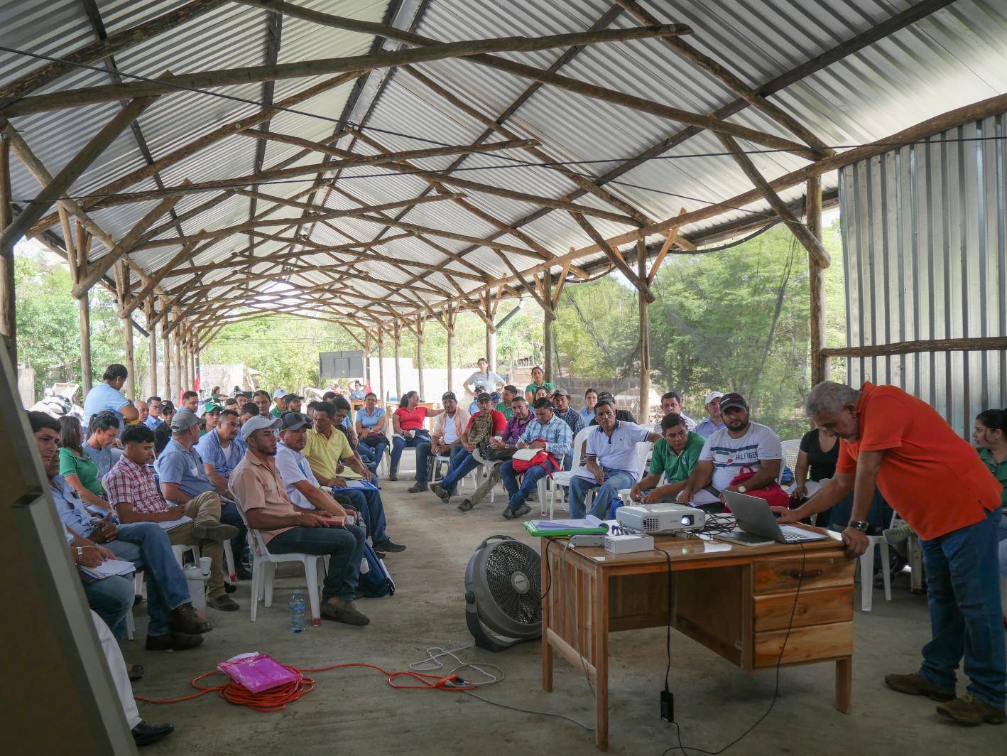 A group of people seated in a rustic covered outdoor space, attentively listening to a speaker at a desk with a laptop and projector.