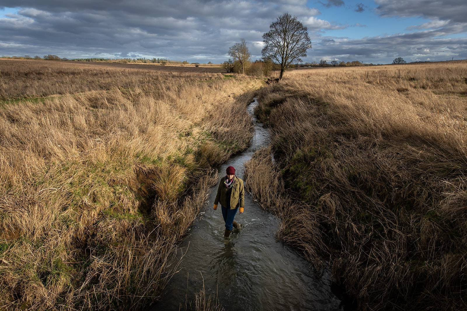A person wearing a red hat and green jacket walks in a narrow stream through an open field under a cloudy sky.