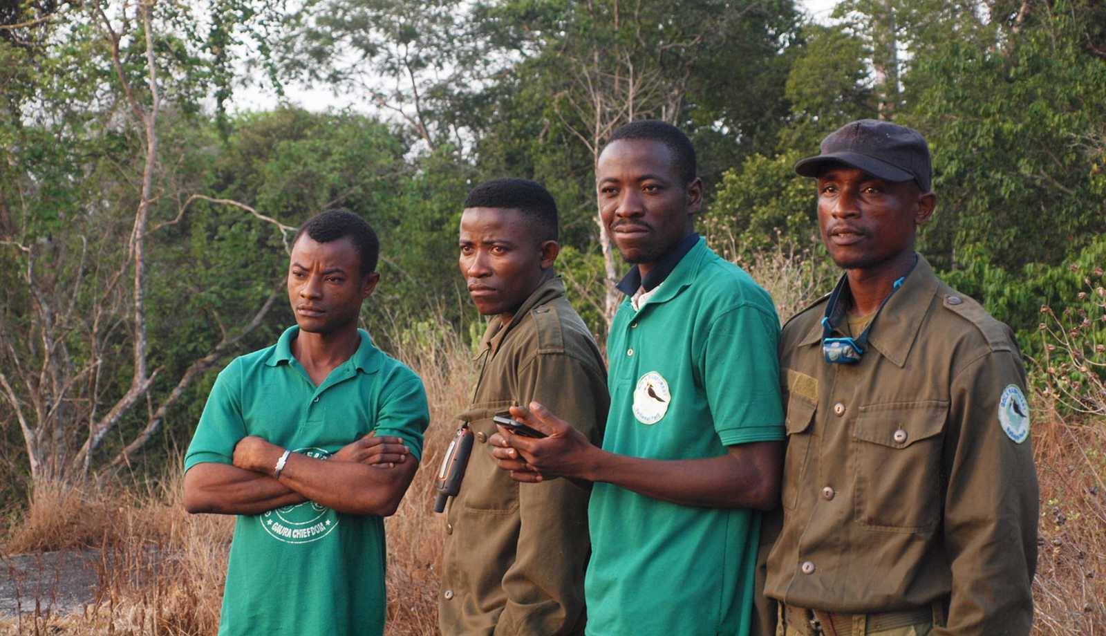 Four men wearing green shirts and jackets stand outdoors, looking serious. The background is a forested area.