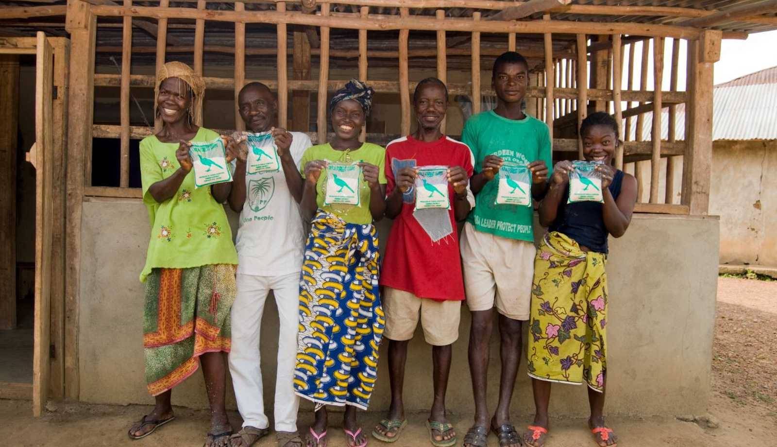 Six people smiling, standing in front of a building, holding up packaged products with blue and green designs.