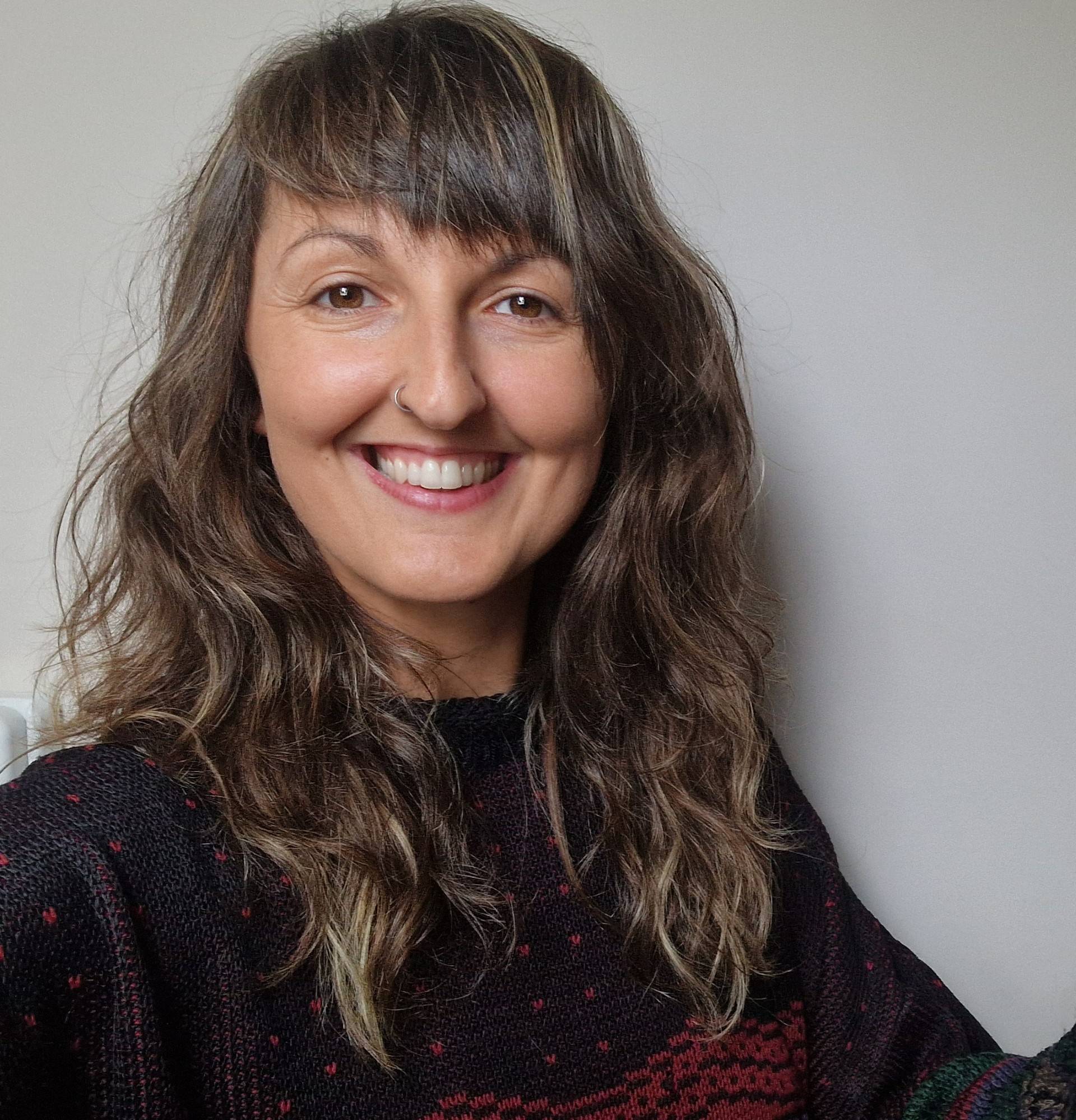 Smiling person with long wavy hair and a nose ring, wearing a patterned black sweater, standing against a plain background.