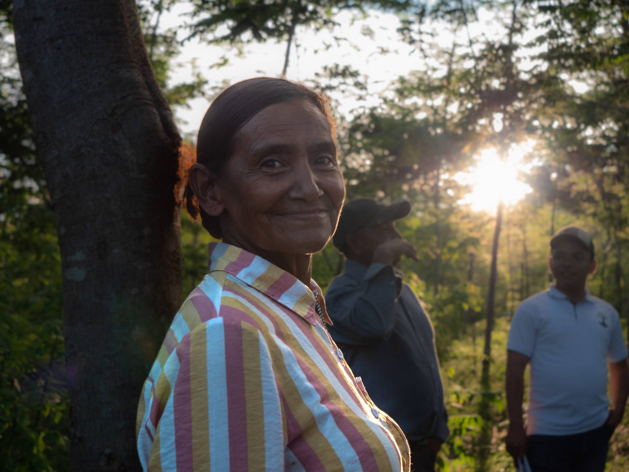 A woman in a striped shirt smiles in a sunlit forest, with two people in the background and sunlight filtering through the trees.