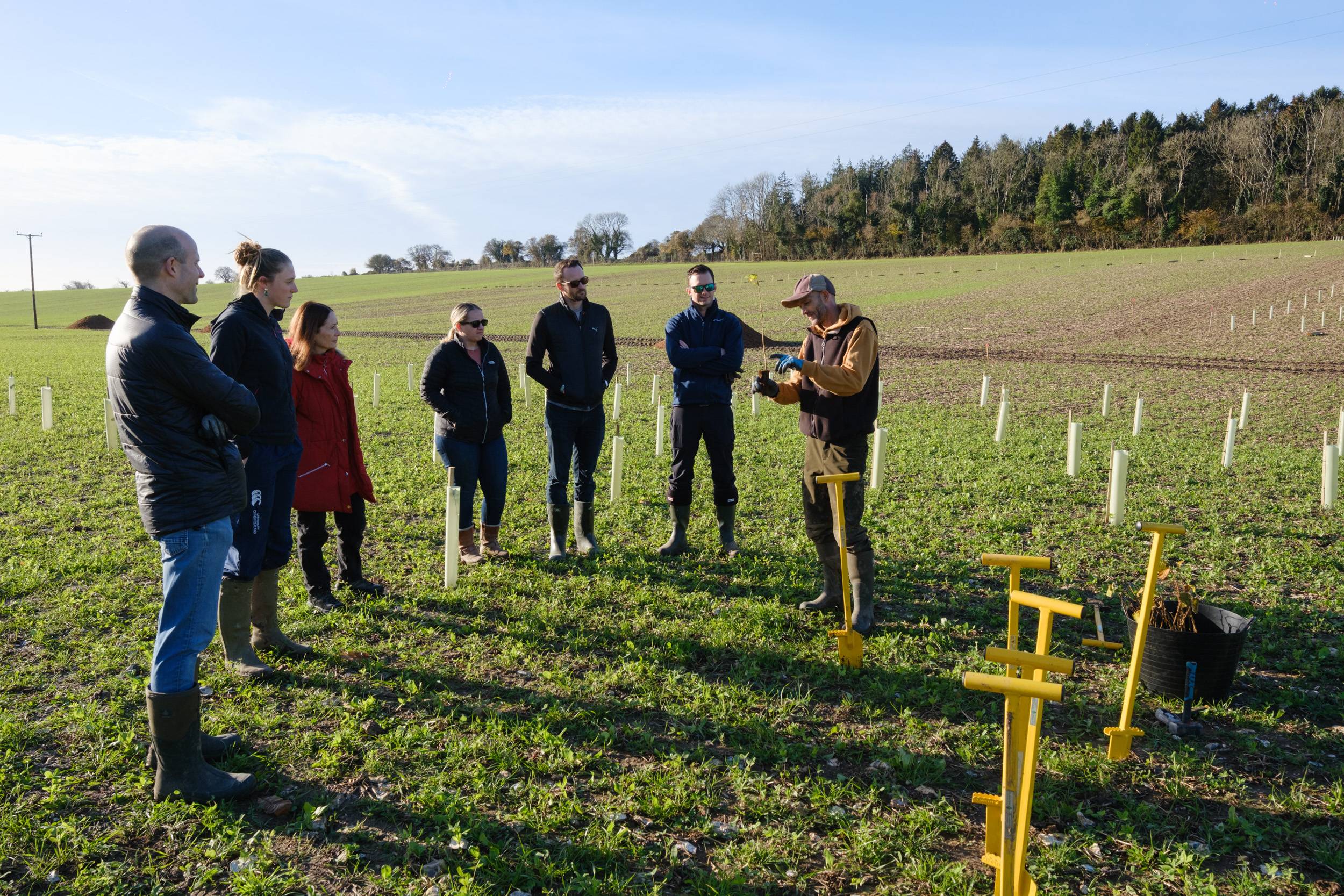 A group of people standing in a field, listening to a person demonstrating with equipment on a sunny day.